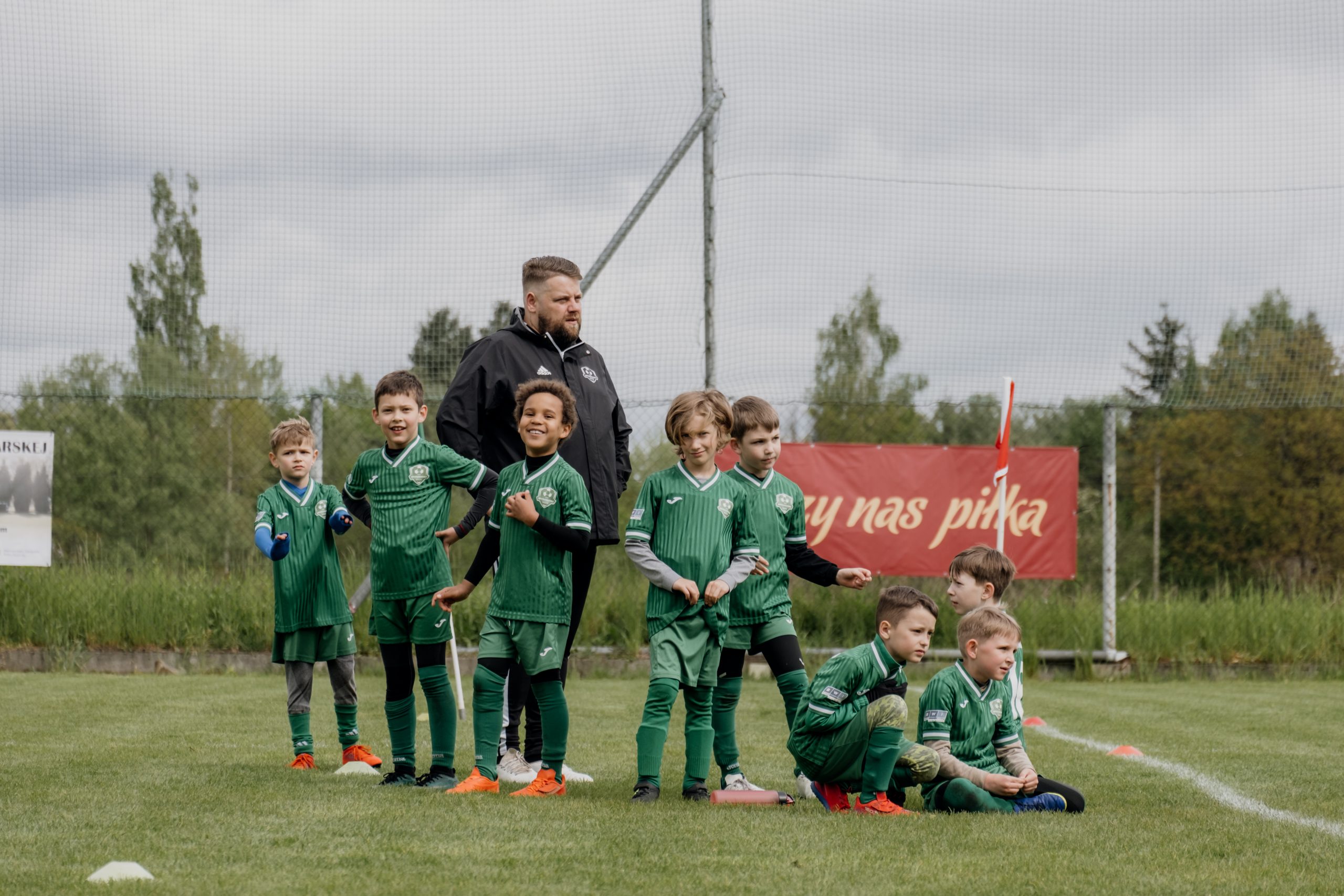 Na zdjęciu widzimy młodych zawodników Opatkowianki w zielonych strojach klubowych, stojących i siedzących w grupie przy linii bocznej boiska. Towarzyszy im trener, który z zaangażowaniem czuwa nad drużyną. Część chłopców uśmiecha się szeroko, inni koncentrują się przed dalszą częścią gry – widać radość, energię i ducha rywalizacji.