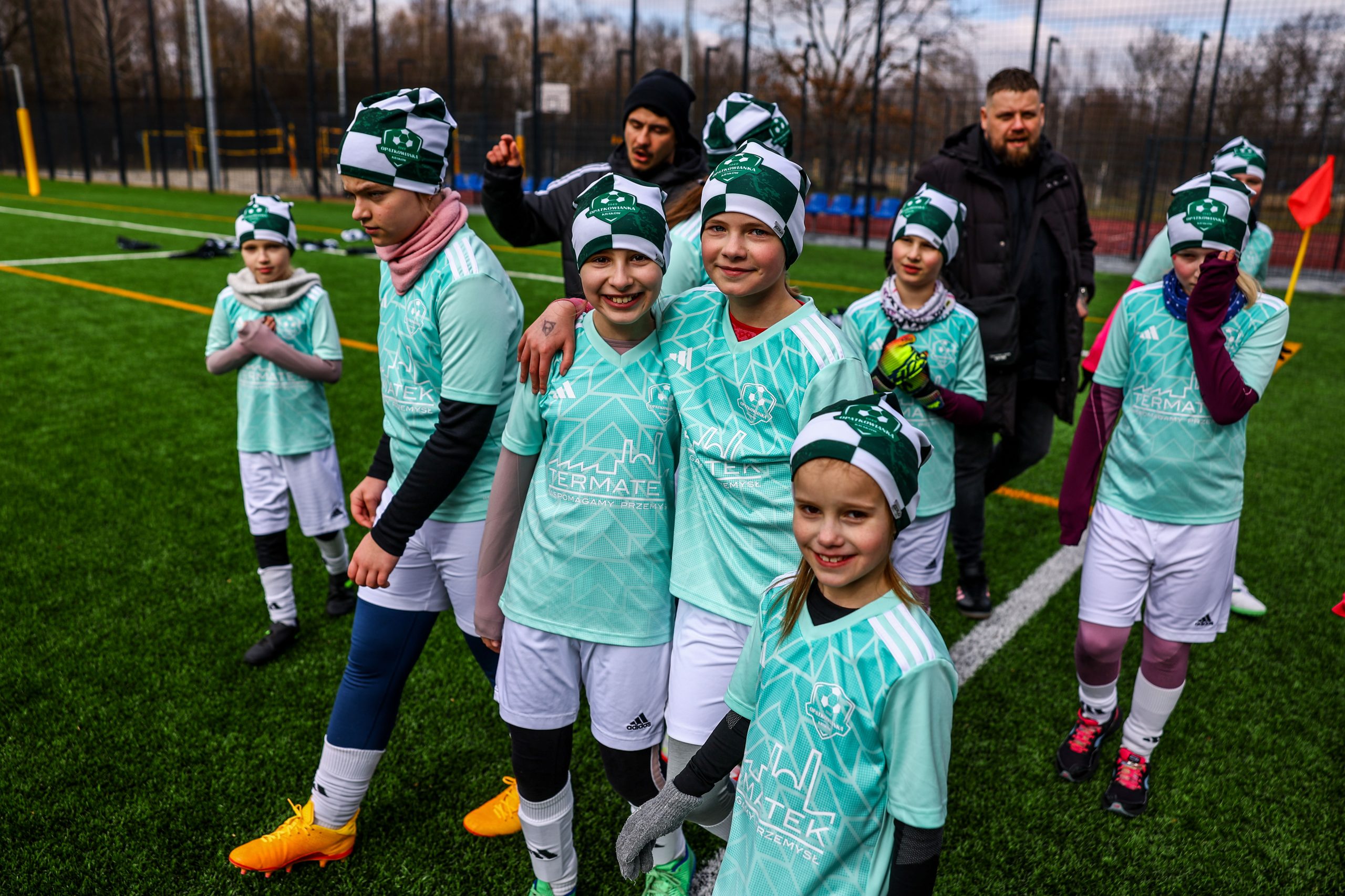 Obrazek przedstawia grupę młodych zawodniczek w jasnozielonych strojach sportowych z herbem „Opatkowianka Kraków”. Dziewczynki uśmiechają się i stoją razem na boisku, niektóre w czapkach klubowych. W tle widać trenerów i osoby towarzyszące. Atmosfera jest radosna i drużynowa.