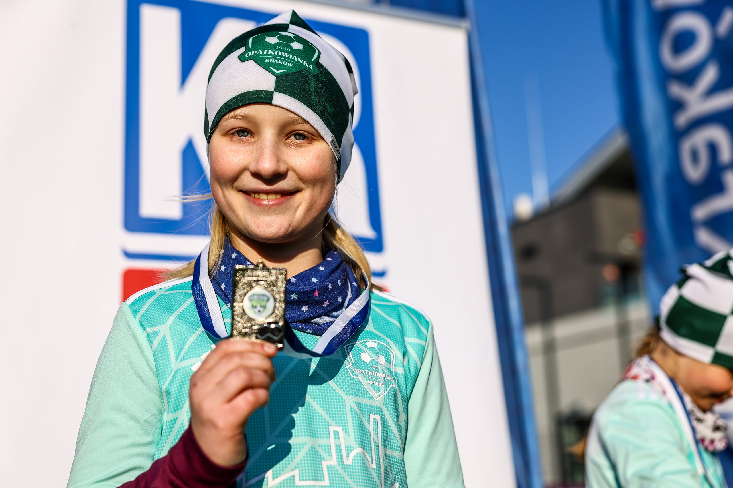 Obrazek przedstawia uśmiechniętą dziewczynkę w czapce i stroju sportowym z herbem „Opatkowianka Kraków”, trzymającą w dłoni medal z tym samym logo. Zdjęcie zostało wykonane podczas wydarzenia sportowego na świeżym powietrzu.